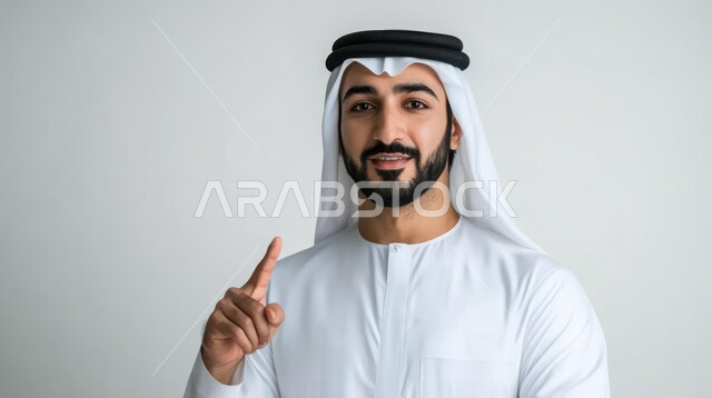 الإشارة و التوضيح إلى شيء ما, تحديد موقع معين, الدلالة على مكان عنصر, بورتريه لشاب عربي خليجي سعودي يرتدي الثوب التقليدي و الشماغ ينظر إلى الكاميرا بإيماءات السعادة و السرور و يشير بإصبع السبابة للأعلى, خلفية رمادية