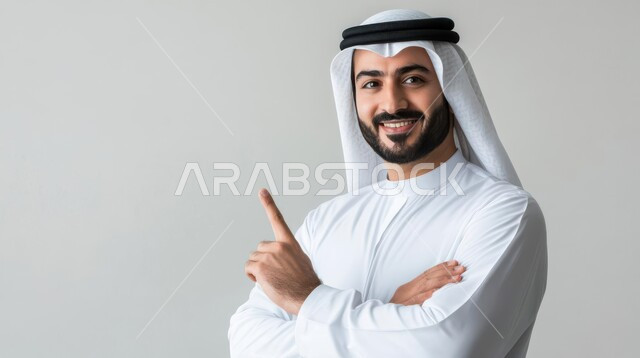 الدلالة على مكان عنصر, تحديد موقع معين, الإشارة و التوضيح إلى شيء ما, بورتريه مقرب لشاب عربي خليجي سعودي يرتدي الثوب التقليدي و الشماغ ينظر إلى الكاميرا بإيماءات السعادة و السرور و يشير بإصبع السبابة للأعلى, خلفية رمادية