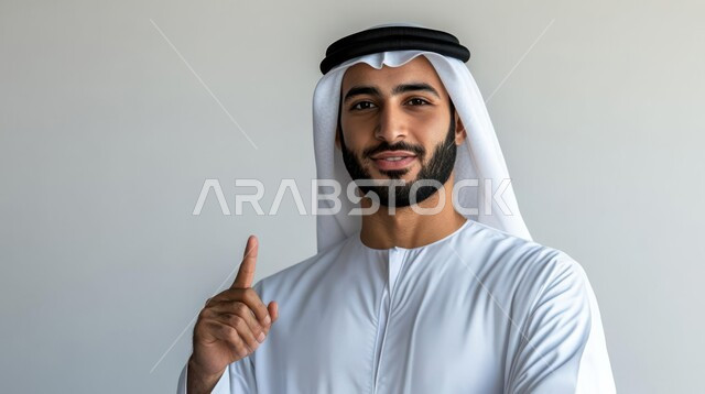 Pointing and explaining something, specifying a specific location, indicating the location of an item, portrait of a young Saudi Arabian Gulf man wearing a traditional thobe and shemagh looking at the camera with gestures of happiness and pleasure and pointing his index finger upwards, gray background