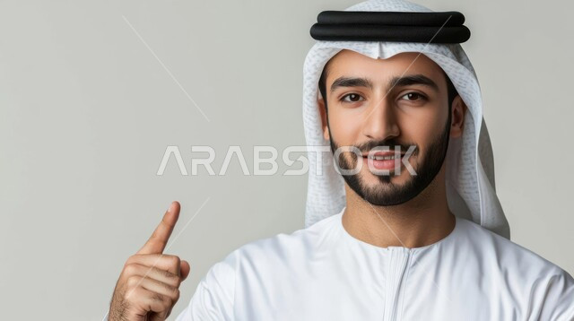 Pointing and explaining something, specifying a specific location, indicating the location of an item, portrait of a young Saudi Arabian Gulf man wearing a traditional thobe and shemagh looking at the camera with gestures of happiness and pleasure and pointing his index finger upwards, gray background