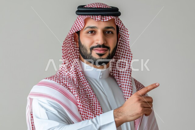 تحديد موقع معين, الإشارة و التوضيح إلى شيء ما, الدلالة على مكان عنصر, بورتريه لشاب عربي خليجي سعودي يرتدي الثوب التقليدي و الشماغ ينظر إلى الكاميرا بإيماءات السعادة و السرور و يشير بإصبع السبابة, خلفية رمادية