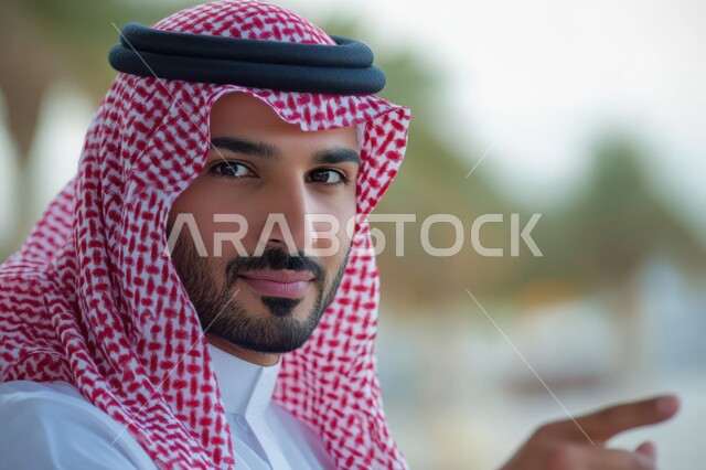 الإشارة و التوضيح إلى شيء ما, تحديد موقع معين, الدلالة على مكان عنصر, صورة مقربة لشاب عربي خليجي سعودي يرتدي الثوب التقليدي و الشماغ ينظر إلى الكاميرا بإيماءات السعادة و السرور و يشير بإصبع السبابة