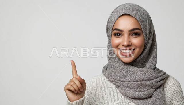تحديد موقع معين, الإشارة و التوضيح إلى شيء ما, الدلالة على مكان عنصر, بورتريه مقرب لإمرأة عربية خليجي سعودية مبتسمة محجبة ترتدي الزي الكاجوال تنظر إلى الكاميرا بإيماءات السعادة والسرور وتشير بإصبع السبابة للأعلى, خلفية رمادية