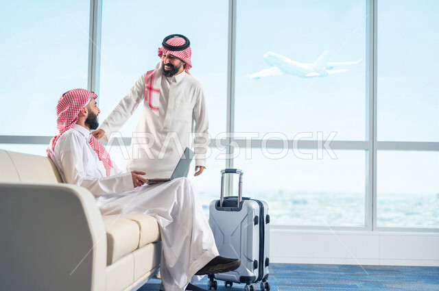 رجل اعمال عربي سعودي  خليجي يلتقي بزميل العمل اثناء الجلوس في صالة الانتظار بالمطار، انجاز العمل من خلال جهاز الحاسوب المحمول،  الاستعداد للسفر في رحلة عمل، قاعة الانتظار في المطار، سياحة وسفر