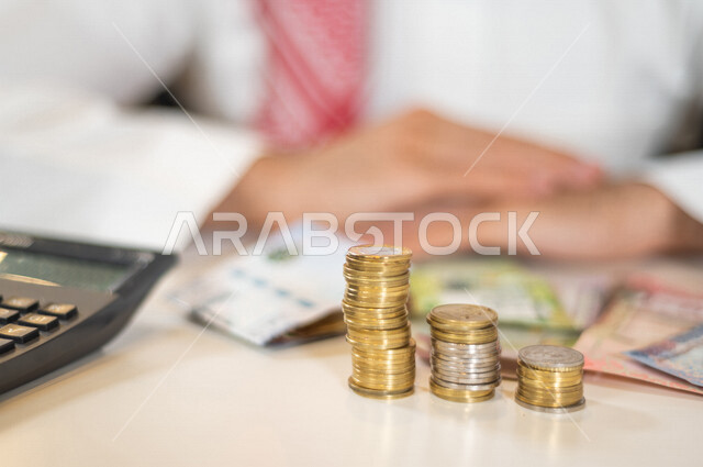 صورة ضبابية لرجل عربي خليجي سعودي ، يعمل على ادارة الحسابات، النمو والأرباح المالية، إدخار وتوفير الاموال، عملات نقدية معدنية وورقية للريال السعودي، استخدام الالة الحاسبة لاجراء العمليات الحسابية