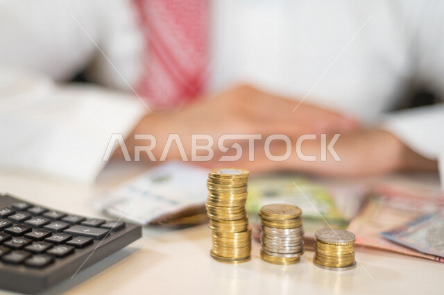 صورة ضبابية لرجل عربي خليجي سعودي ، يعمل على ادارة الحسابات، النمو والأرباح المالية، إدخار وتوفير الاموال، عملات نقدية معدنية وورقية للريال السعودي، استخدام الالة الحاسبة لاجراء العمليات الحسابية