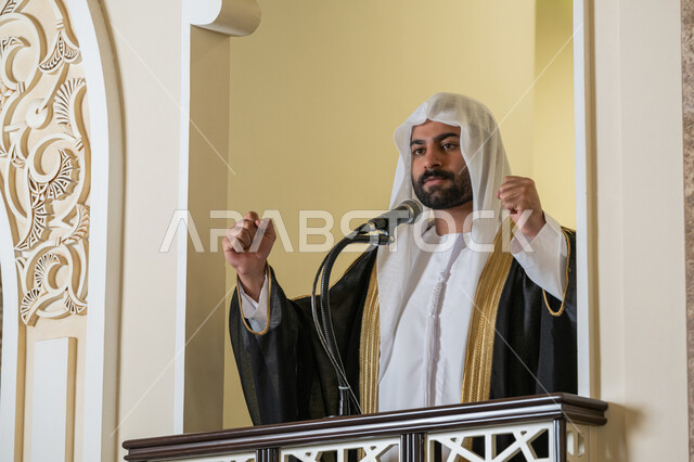 مفهوم العبادة والتقرب إلى الله تعالى, تعزيز الروحانية الجماعية, توجيه الناس نحو الحق والهداية, دور أئمة المساجد والخطباء في نشر الدين، إمام عربي خليجي إماراتي يرتدي البشت والكندورة والغترة البيضاء يقف أمام مكبر الصوت ويلقي الخطبة على المنبر