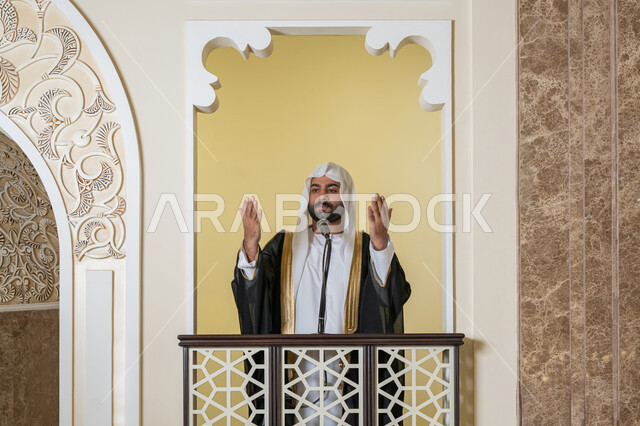 مفهوم الخشوع والتضرع, طلب الرحمة والمغفرة, إمامة المسلمين في المسجد، العبادة والتقرب إلى الله، تعزيز الروحانية الجماعية, صورة لامام عربي خليجي إماراتي يرتدي البشت والكندورة والغترة البيضاء يقف على المنبر ويرفع يديه بتعابير الدعاء
