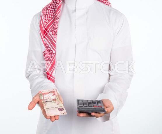 بورتريه لرجل عربي خليجي سعودي، يحمل في يده عملة ورقية والة حاسبة في اليد الاخرى، العملات الورقية للريال السعودية، استخدام الالة الحاسبة لاجراء العمليات الحسابية، ادارة الحسابات