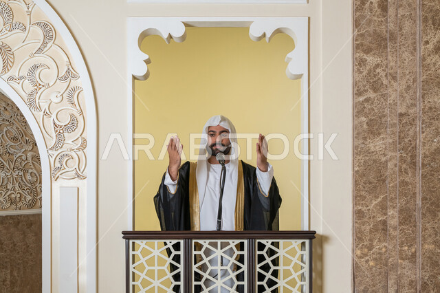 تعزيز الروحانية الجماعية, إمامة المسلمين في المسجد، مفهوم الخشوع والتضرع, طلب الرحمة والمغفرة, العبادة والتقرب إلى الله،  صورة مقربة لامام عربي خليجي إماراتي يرتدي البشت والكندورة والغترة البيضاء يقف على المنبر ويرفع يديه بتعابير الدعاء
