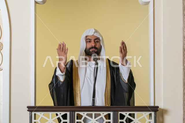 إمامة المسلمين في المسجد، مفهوم الخشوع والتضرع, طلب الرحمة والمغفرة, العبادة والتقرب إلى الله، تعزيز الروحانية الجماعية, صورة مقربة لامام عربي خليجي إماراتي يرتدي البشت والكندورة والغترة البيضاء يقف على المنبر ويرفع يديه بتعابير الدعاء