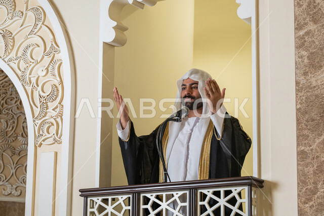 تعزيز الروحانية الجماعية, إمامة المسلمين في المسجد، مفهوم الخشوع والتضرع, طلب الرحمة والمغفرة, العبادة والتقرب إلى الله،  صورة مقربة لامام عربي خليجي إماراتي يرتدي البشت والكندورة والغترة البيضاء يقف على المنبر ويرفع يديه بتعابير الدعاء