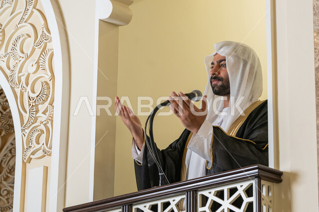 مفهوم الخشوع والتضرع, طلب الرحمة والمغفرة, إمامة المسلمين في المسجد، العبادة والتقرب إلى الله، تعزيز الروحانية الجماعية, صورة لامام عربي خليجي إماراتي يرتدي البشت والكندورة والغترة البيضاء يقف على المنبر ويرفع يديه بتعابير الدعاء