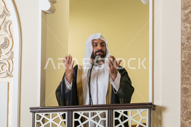إمامة المسلمين في المسجد، مفهوم الخشوع والتضرع, طلب الرحمة والمغفرة, العبادة والتقرب إلى الله، تعزيز الروحانية الجماعية, صورة مقربة لامام عربي خليجي إماراتي يرتدي البشت والكندورة والغترة البيضاء يقف على المنبر ويرفع يديه بتعابير الدعاء