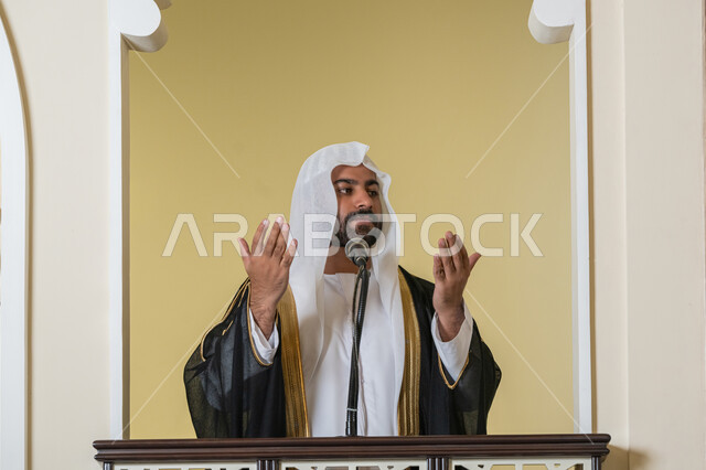مفهوم الخشوع والتضرع, طلب الرحمة والمغفرة, إمامة المسلمين في المسجد، العبادة والتقرب إلى الله، تعزيز الروحانية الجماعية, صورة لامام عربي خليجي إماراتي يرتدي البشت والكندورة والغترة البيضاء يقف على المنبر ويرفع يديه بتعابير الدعاء