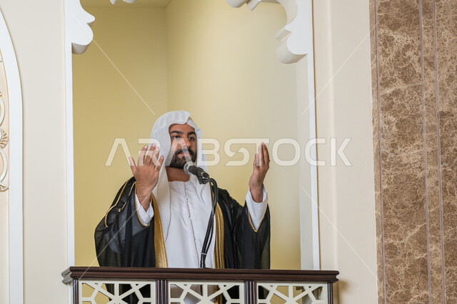 تعزيز الروحانية الجماعية, إمامة المسلمين في المسجد، مفهوم الخشوع والتضرع, طلب الرحمة والمغفرة, العبادة والتقرب إلى الله،  صورة مقربة لامام عربي خليجي إماراتي يرتدي البشت والكندورة والغترة البيضاء يقف على المنبر ويرفع يديه بتعابير الدعاء