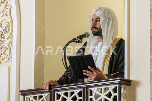 توجيه الناس لطريق الحق والهداية, الحث على أداء العبادات والفرائض الدينية, إلقاء خطاب و دروس توجيهية على المسلمين, إمام مسجد عربي خليجي إماراتي يرتدي البشت والكندورة والغترة البيضاء يقف على المنبر ويلقي خطبة يوم الجمعة من الجهاز اللوحي