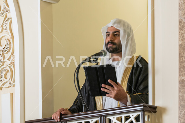 الحث على أداء العبادات والفرائض الدينية, إلقاء خطاب و دروس توجيهية على المسلمين, توجيه الناس لطريق الحق والهداية, إمام مسجد عربي خليجي إماراتي يرتدي البشت والكندورة والغترة البيضاء يقف على المنبر ويلقي خطبة يوم الجمعة من الجهاز اللوحي