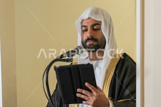 الحث على أداء العبادات والفرائض الدينية, إلقاء خطاب و دروس توجيهية على المسلمين, توجيه الناس لطريق الحق والهداية, إمام مسجد عربي خليجي إماراتي يرتدي البشت والكندورة والغترة البيضاء يقف على المنبر ويلقي خطبة يوم الجمعة من الجهاز اللوحي