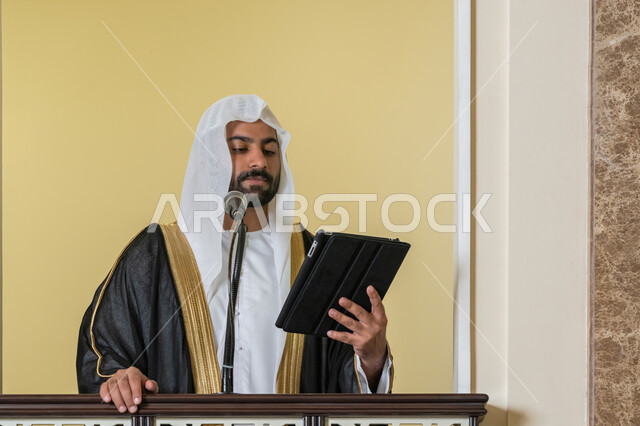 إلقاء خطاب و دروس توجيهية على المسلمين, توجيه الناس لطريق الحق والهداية, الحث على أداء العبادات والفرائض الدينية, إمام مسجد عربي خليجي إماراتي يرتدي البشت والكندورة والغترة البيضاء يقف على المنبر ويلقي خطبة يوم الجمعة من الجهاز اللوحي