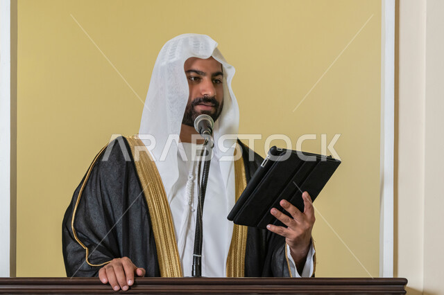 توجيه الناس لطريق الحق والهداية, الحث على أداء العبادات والفرائض الدينية, إلقاء خطاب و دروس توجيهية على المسلمين, إمام مسجد عربي خليجي إماراتي يرتدي البشت والكندورة والغترة البيضاء يقف على المنبر ويلقي خطبة يوم الجمعة من الجهاز اللوحي