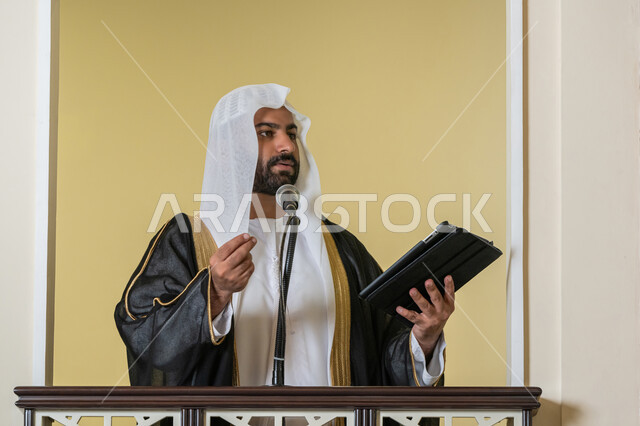 الحث على أداء العبادات والفرائض الدينية, إلقاء خطاب و دروس توجيهية على المسلمين, توجيه الناس لطريق الحق والهداية, إمام مسجد عربي خليجي إماراتي يرتدي البشت والكندورة والغترة البيضاء يقف على المنبر ويلقي خطبة يوم الجمعة من الجهاز اللوحي