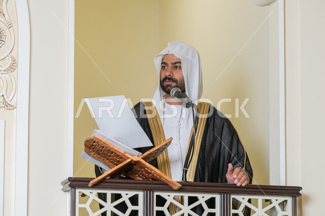 حث المسلمين لأداء الفرائض, توجيه الناس نحو الحق والهداية, مفهوم العبادة والتقرب إلى الله تعالى, دور أئمة المساجد في نشر الدين، صورة مقربة لإمام مسجد عربي خليجي إماراتي يرتدي البشت والكندورة والغترة البيضاء يقف على المنبر يمسك بيده أوراق ويلقي خطبة يوم الجمعة