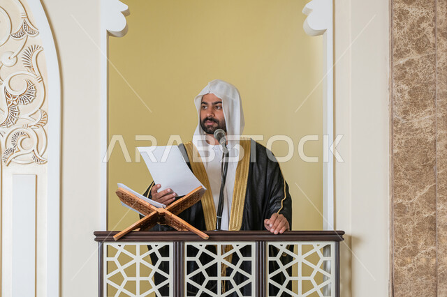 مفهوم العبادة والتقرب إلى الله تعالى, حث المسلمين لأداء الفرائض, توجيه الناس نحو الحق والهداية, دور أئمة المساجد في نشر الدين، صورة مقربة لإمام مسجد عربي خليجي إماراتي يرتدي البشت والكندورة والغترة البيضاء يقف على المنبر يمسك بيده أوراق ويلقي خطبة يوم الجمعة