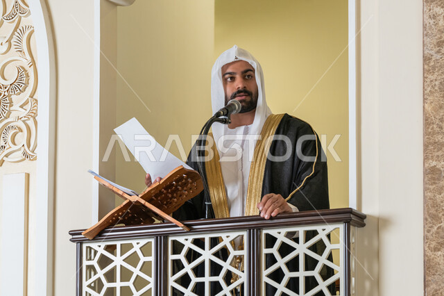حث المسلمين لأداء الفرائض, توجيه الناس نحو الحق والهداية, مفهوم العبادة والتقرب إلى الله تعالى, دور أئمة المساجد في نشر الدين، صورة مقربة لإمام مسجد عربي خليجي إماراتي يرتدي البشت والكندورة والغترة البيضاء يقف على المنبر يمسك بيده أوراق ويلقي خطبة يوم الجمعة