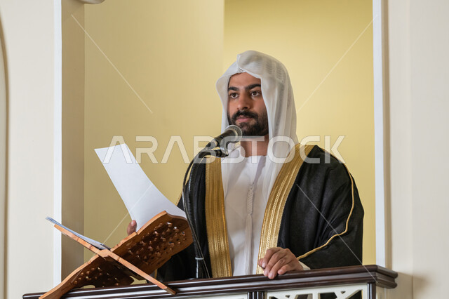 توجيه الناس نحو الحق والهداية, حث المسلمين لأداء الفرائض, مفهوم العبادة والتقرب إلى الله تعالى, دور أئمة المساجد في نشر الدين، صورة مقربة لإمام مسجد عربي خليجي إماراتي يرتدي البشت والكندورة والغترة البيضاء يقف على المنبر يمسك بيده أوراق ويلقي خطبة يوم الجمعة