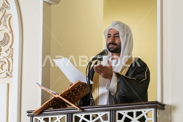 مفهوم العبادة والتقرب إلى الله تعالى, حث المسلمين لأداء الفرائض, توجيه الناس نحو الحق والهداية, دور أئمة المساجد في نشر الدين، صورة مقربة لإمام مسجد عربي خليجي إماراتي يرتدي البشت والكندورة والغترة البيضاء يقف على المنبر يمسك بيده أوراق ويلقي خطبة يوم الجمعة