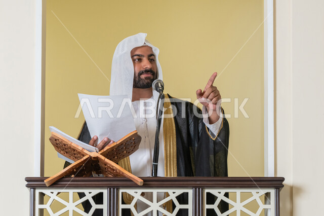 توجيه الناس نحو الحق والهداية, حث المسلمين لأداء الفرائض, مفهوم العبادة والتقرب إلى الله تعالى, دور أئمة المساجد في نشر الدين، صورة مقربة لإمام مسجد عربي خليجي إماراتي يرتدي البشت والكندورة والغترة البيضاء يقف على المنبر يمسك بيده أوراق ويلقي خطبة يوم الجمعة