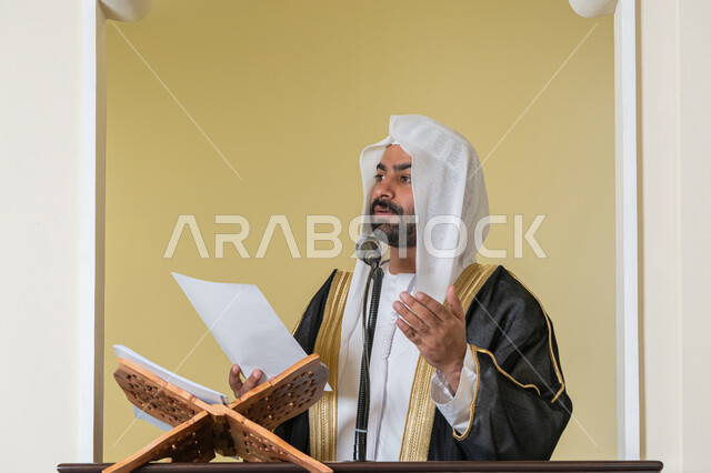 حث المسلمين لأداء الفرائض, توجيه الناس نحو الحق والهداية, مفهوم العبادة والتقرب إلى الله تعالى, دور أئمة المساجد في نشر الدين، صورة مقربة لإمام مسجد عربي خليجي إماراتي يرتدي البشت والكندورة والغترة البيضاء يقف على المنبر يمسك بيده أوراق ويلقي خطبة يوم الجمعة