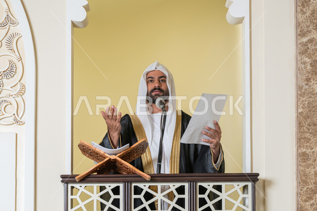 مفهوم العبادة والتقرب إلى الله تعالى, حث المسلمين لأداء الفرائض, توجيه الناس نحو الحق والهداية, دور أئمة المساجد في نشر الدين، صورة مقربة لإمام مسجد عربي خليجي إماراتي يرتدي البشت والكندورة والغترة البيضاء يقف على المنبر يمسك بيده أوراق ويلقي خطبة يوم الجمعة
