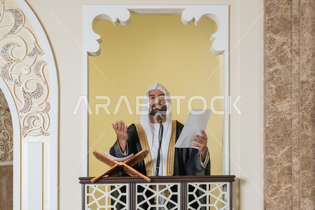 Directing people towards truth and guidance, urging Muslims to perform religious duties, the concept of worship and getting closer to God Almighty, the role of imams of mosques in spreading religion, a close-up photo of the imam of an Arab Gulf-Emirati mosque wearing a bisht, kandura, and white ghutra, standing on the pulpit holding papers in his hand and delivering a Friday sermon