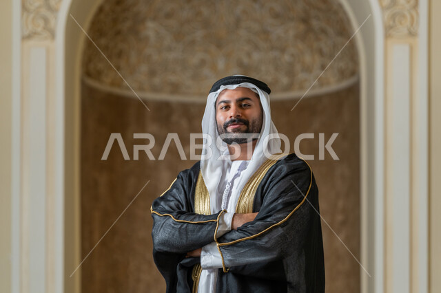 مفهوم الأناقة و الرجولة, الاهتمام بالمظهر الخارجي, صورة مقربة لرجل عربي خليجي اماراتي مبتسم يرتدي البشت الاسود و الغترة يقف بايماءات ايدي متقاطعة، النظر إلى الكاميرا بتعابير السعادة و السرور