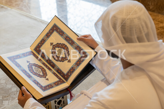 التعبد في بيوت الرحمن، تدبر الآيات القرآنية والتفكر بها, مفهوم العبادة والطاعة والتقرب الى الله, الالتزام بالورد اليومي,  صورة من الخلف لرجل عربي خليجي إماراتي يرتدي الكندورة والغترة البيضاء يجلس في المسجد ويقرأ من المصحف الشريف، التمعن بسور القرآن الكريم