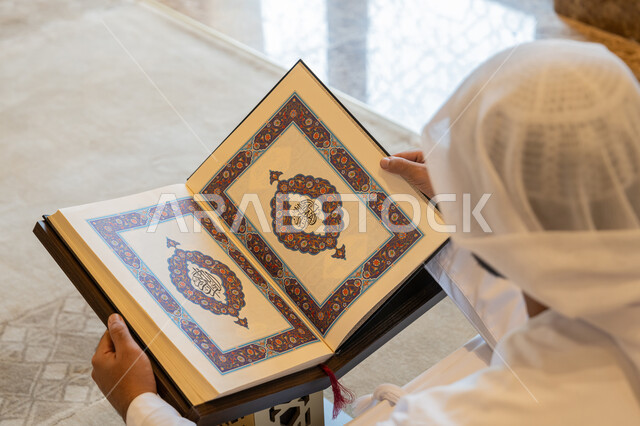تدبر الآيات القرآنية والتفكر بها، التمعن بسور القرآن الكريم, مفهوم العبادة والطاعة والتقرب الى الله, الالتزام بالورد اليومي,  صورة من الخلف لرجل عربي خليجي إماراتي يرتدي الكندورة والغترة البيضاء يجلس في المسجد ويقرأ من المصحف الشريف, التعبد في بيوت الرحمن