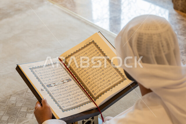 التعبد في بيوت الرحمن، تدبر الآيات القرآنية والتفكر بها, مفهوم العبادة والطاعة والتقرب الى الله, الالتزام بالورد اليومي,  صورة من الخلف لرجل عربي خليجي إماراتي يرتدي الكندورة والغترة البيضاء يجلس في المسجد ويقرأ من المصحف الشريف، التمعن بسور القرآن الكريم