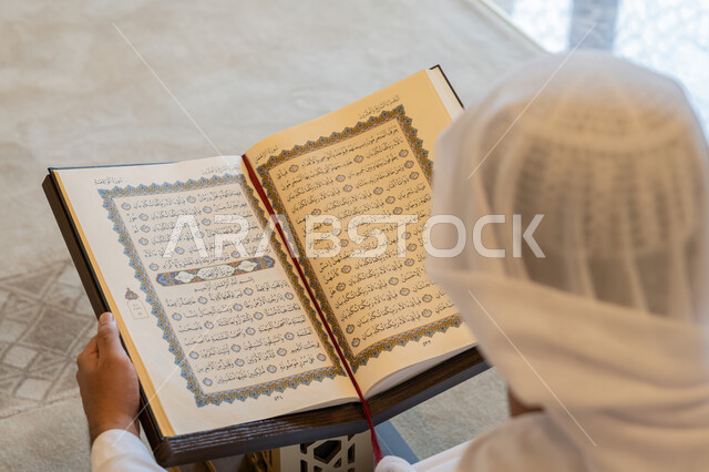 تدبر الآيات القرآنية والتفكر بها، التمعن بسور القرآن الكريم, مفهوم العبادة والطاعة والتقرب الى الله, الالتزام بالورد اليومي,  صورة من الخلف لرجل عربي خليجي إماراتي يرتدي الكندورة والغترة البيضاء يجلس في المسجد ويقرأ من المصحف الشريف, التعبد في بيوت الرحمن