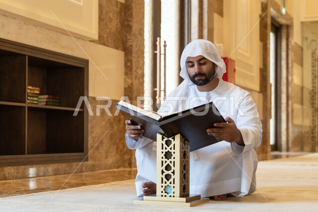 التمعن بسور القرآن الكريم, مفهوم العبادة والطاعة والتقرب الى الله, تدبر الآيات القرآنية والتفكر بها، الالتزام بالورد اليومي, رجل عربي خليجي إماراتي يرتدي الكندورة والغترة البيضاء يجلس في المسجد ويقرأ من المصحف الشريف, التعبد في بيوت الرحمن