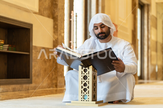 The concept of worship, obedience and drawing closer to God, contemplating the chapters of the Holy Quran, pondering and reflecting on the verses of the Quran, committing to the daily rosary, an Arab Gulf Emirati man wearing a white kandura and ghutra sitting in the mosque and reading from the Holy Quran, worship in the houses of the Most Merciful