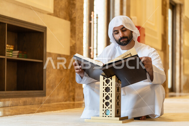 التمعن بسور القرآن الكريم, مفهوم العبادة والطاعة والتقرب الى الله, تدبر الآيات القرآنية والتفكر بها، الالتزام بالورد اليومي, رجل عربي خليجي إماراتي يرتدي الكندورة والغترة البيضاء يجلس في المسجد ويقرأ من المصحف الشريف, التعبد في بيوت الرحمن