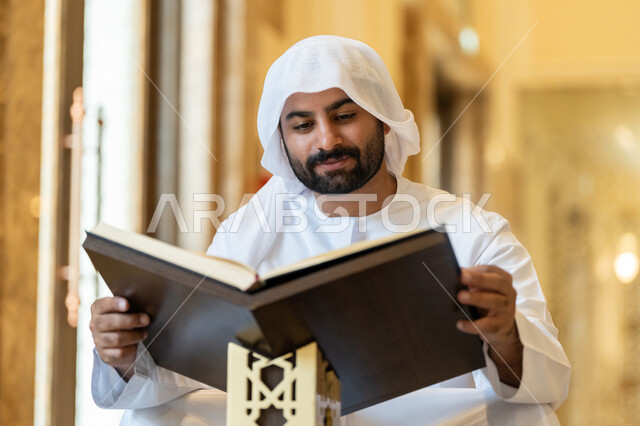 The concept of worship, obedience and drawing closer to God, contemplating the chapters of the Holy Quran, pondering and reflecting on the verses of the Quran, committing to the daily rosary, an Arab Gulf Emirati man wearing a white kandura and ghutra sitting in the mosque and reading from the Holy Quran, worship in the houses of the Most Merciful