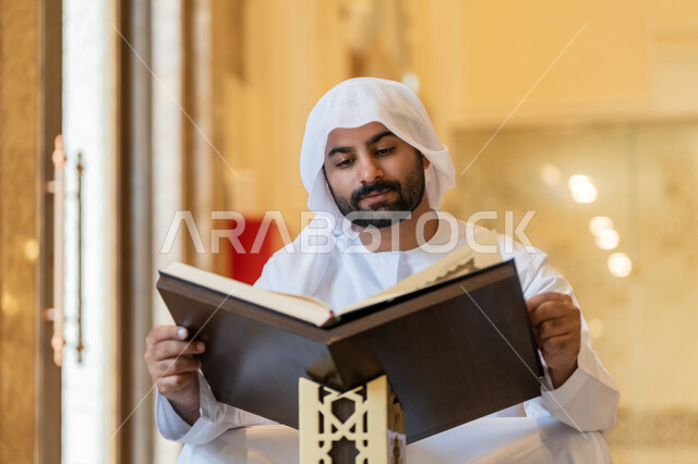 التمعن بسور القرآن الكريم, مفهوم العبادة والطاعة والتقرب الى الله, تدبر الآيات القرآنية والتفكر بها، الالتزام بالورد اليومي, رجل عربي خليجي إماراتي يرتدي الكندورة والغترة البيضاء يجلس في المسجد ويقرأ من المصحف الشريف, التعبد في بيوت الرحمن