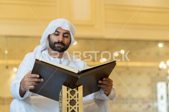 مفهوم العبادة والطاعة والتقرب الى الله, التمعن بسور القرآن الكريم, تدبر الآيات القرآنية والتفكر بها، الالتزام بالورد اليومي, رجل عربي خليجي إماراتي يرتدي الكندورة والغترة البيضاء يجلس في المسجد ويقرأ من المصحف الشريف, التعبد في بيوت الرحمن
