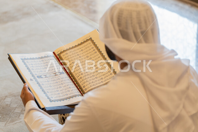 التعبد في بيوت الرحمن، تدبر الآيات القرآنية والتفكر بها, مفهوم العبادة والطاعة والتقرب الى الله, الالتزام بالورد اليومي,  صورة من الخلف لرجل عربي خليجي إماراتي يرتدي الكندورة والغترة البيضاء يجلس في المسجد ويقرأ من المصحف الشريف، التمعن بسور القرآن الكريم