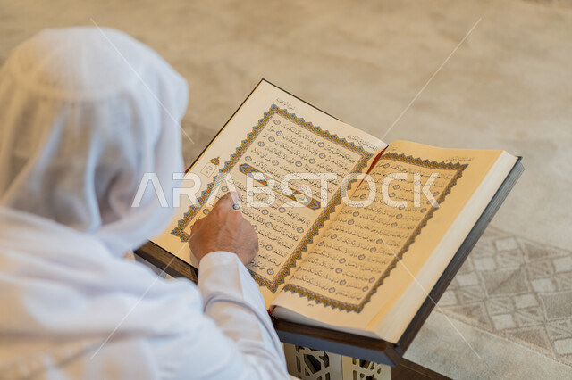 تدبر الآيات القرآنية والتفكر بها، التمعن بسور القرآن الكريم, مفهوم العبادة والطاعة والتقرب الى الله, الالتزام بالورد اليومي,  صورة من الخلف لرجل عربي خليجي إماراتي يرتدي الكندورة والغترة البيضاء يجلس في المسجد ويقرأ من المصحف الشريف, التعبد في بيوت الرحمن
