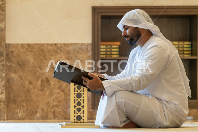 مفهوم العبادة والطاعة والتقرب الى الله, التمعن بسور القرآن الكريم, تدبر الآيات القرآنية والتفكر بها، الالتزام بالورد اليومي, رجل عربي خليجي إماراتي يرتدي الكندورة والغترة البيضاء يجلس في المسجد ويقرأ من المصحف الشريف, التعبد في بيوت الرحمن