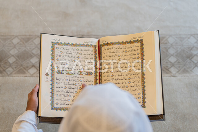 العبادة والطاعة والتقرب الى الله,  تدبر الآيات القرآنية والتفكر بها, الالتزام بالورد اليومي,  صورة من الاعلى لرجل عربي خليجي إماراتي يرتدي الكندورة والغترة البيضاء يجلس في المسجد ويقرأ من المصحف الشريف، التمعن بسور القرآن الكريم، التعبد في بيوت الرحمن